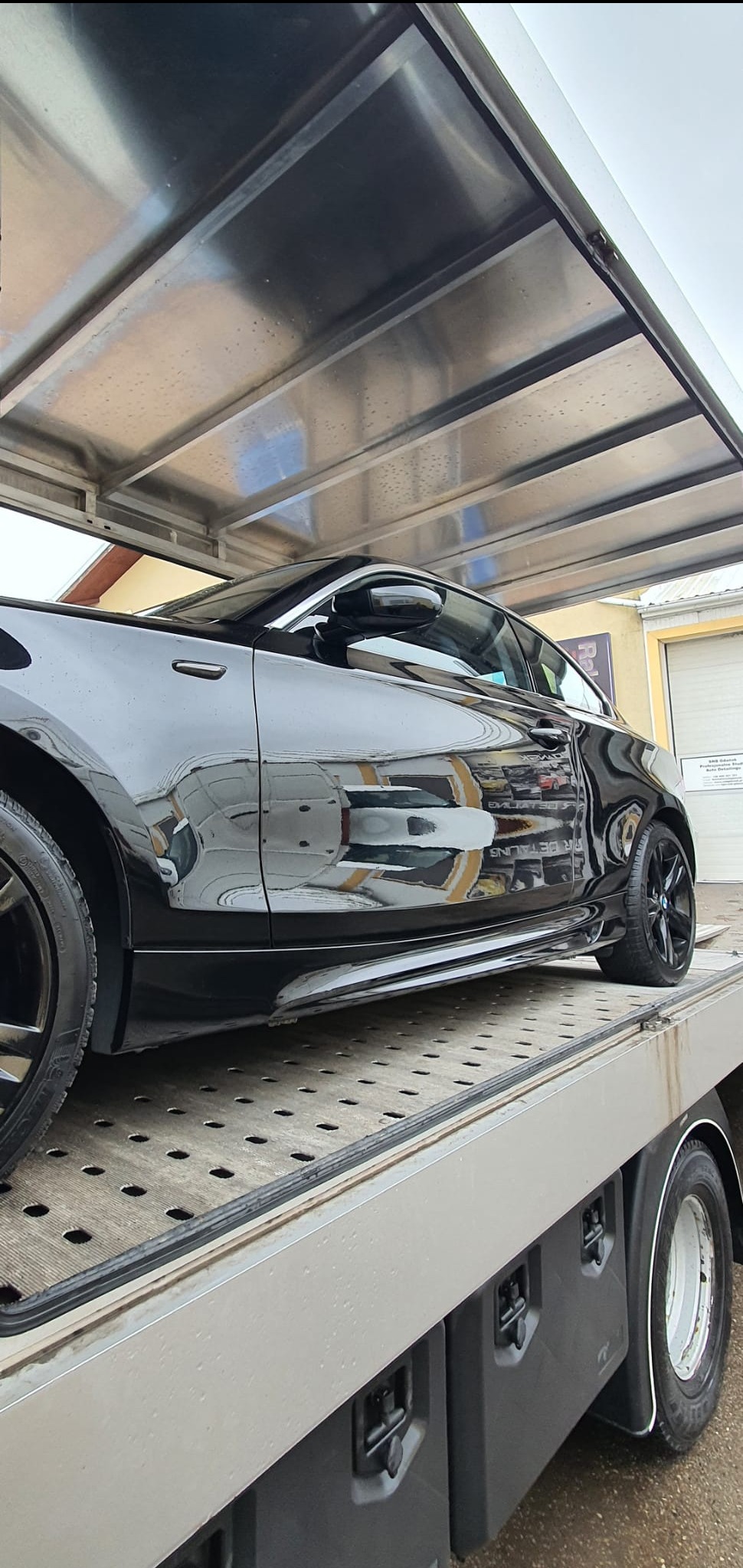 Błyszczący, czarny samochód na lawecie pod zadaszeniem myjni, odbijający otoczenie w lakierze. Widoczny fragment opony i karoserii.