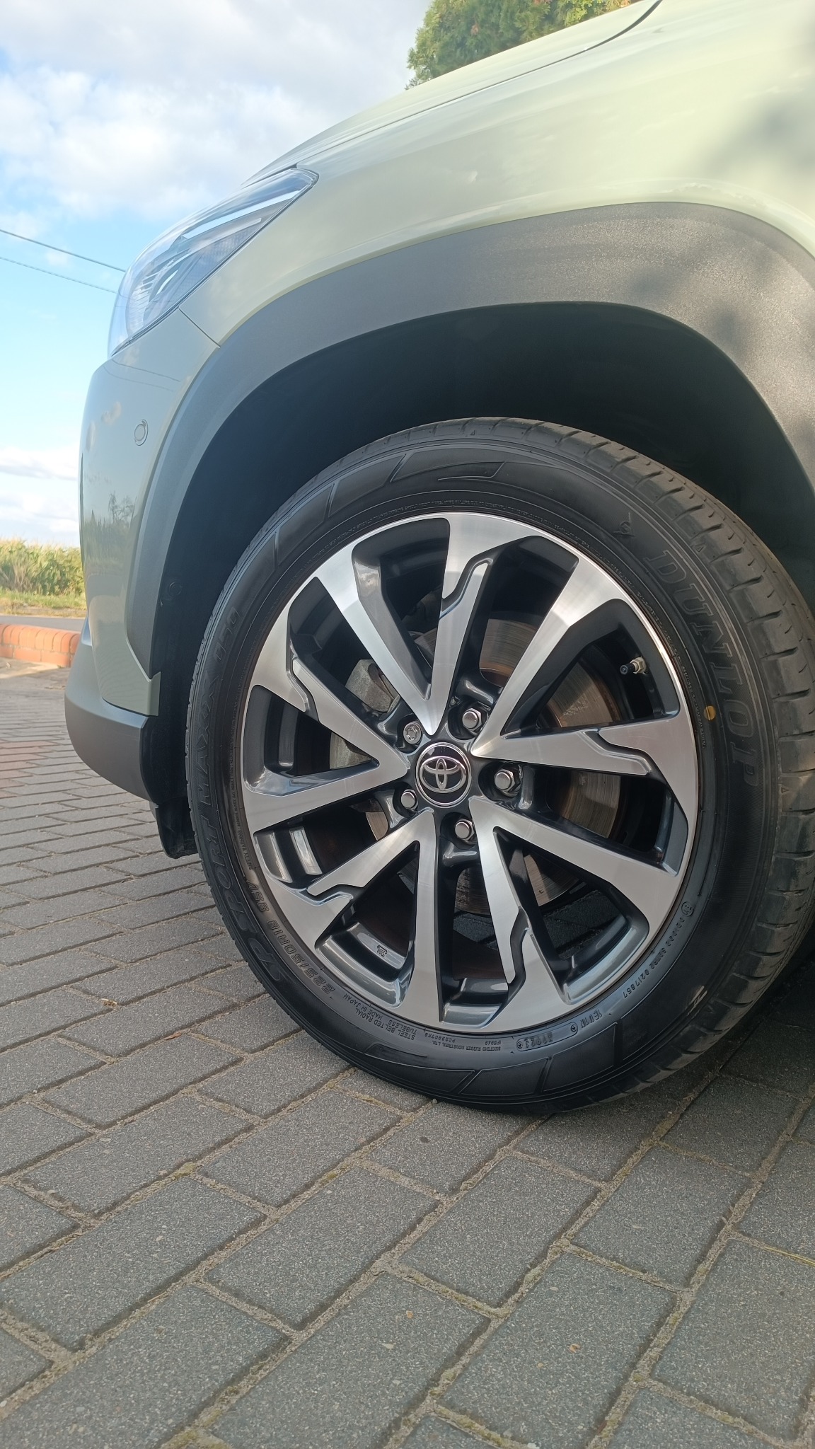 Felga aluminiowa z logo Toyota na oponie Dunlop, widoczna na tle fragmentu karoserii auta w odcieniach zieleni i szarości, zaparkowanego na szarej kostce brukowej.