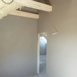 Świeżo wykonane szare ściany w surowym stanie, z widoczną izolacją poddasza i drewnianymi belkami stropowymi, fragment otworu drzwiowego w tle.