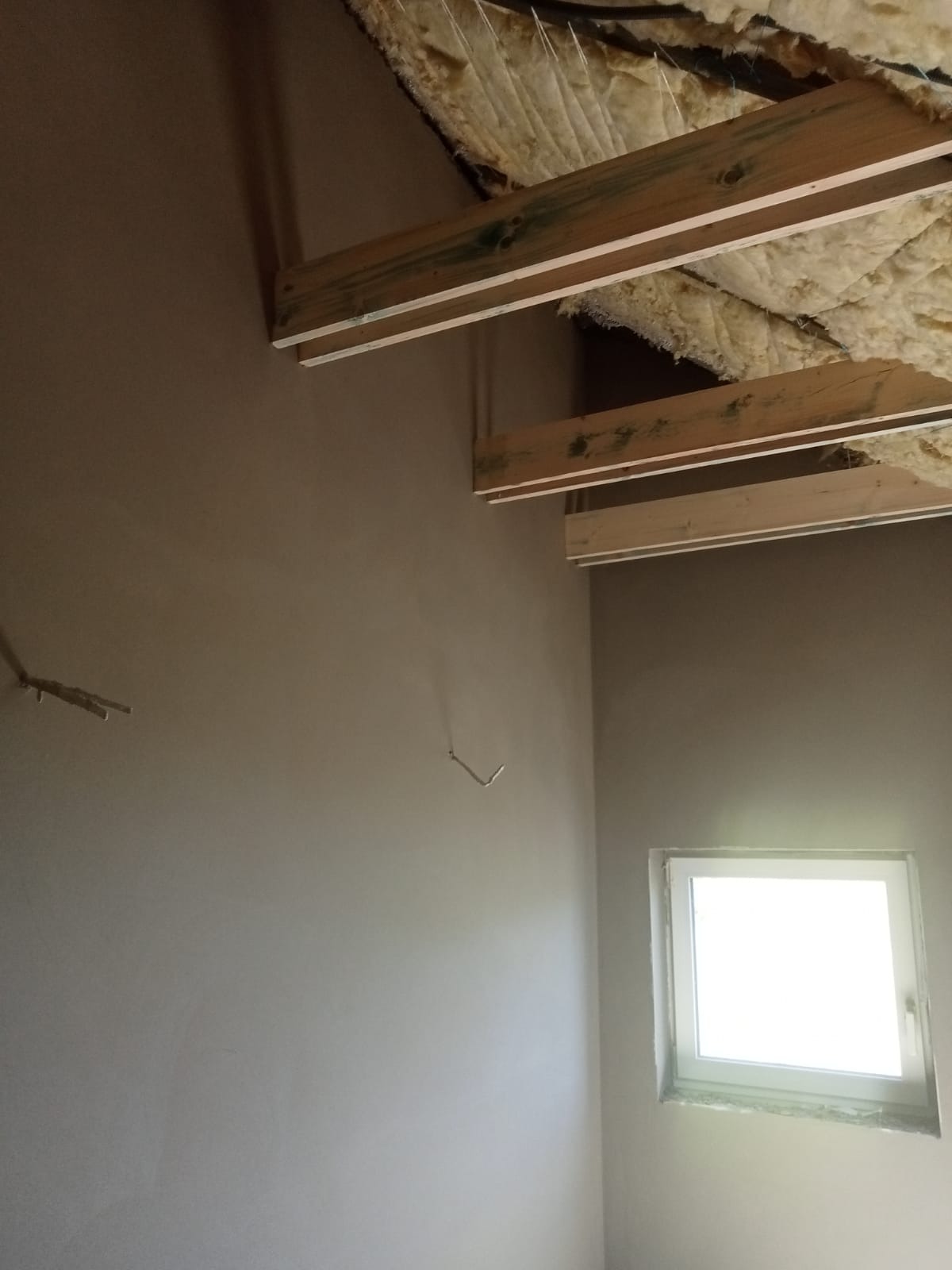 Wnętrze pomieszczenia po nałożeniu szarej wyprawy, widoczne drewniane elementy konstrukcji dachu z izolacją oraz wystające metalowe elementy mocujące.