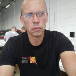 Kostiantyn Mishchenko - Portret mężczyzny w okularach i czarnym T-shircie z logo firmy, z widocznymi pracownikami w tle w jasnym pomieszczeniu.