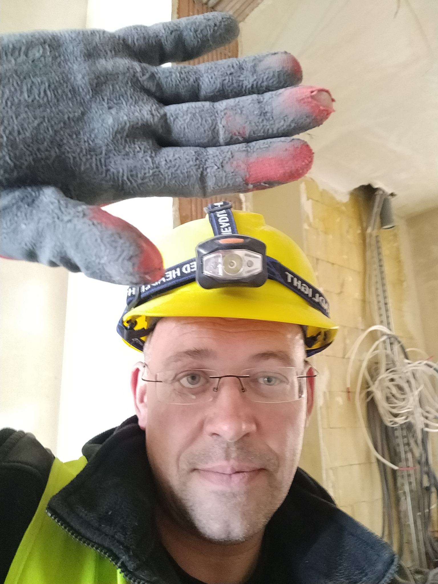 Mężczyzna w okularach i żółtym kasku z latarką, w tle ściana w trakcie remontu z widocznymi kablami i instalacjami, z przodu rękawica robocza z czerwonymi zabrudzeniami na palcach.