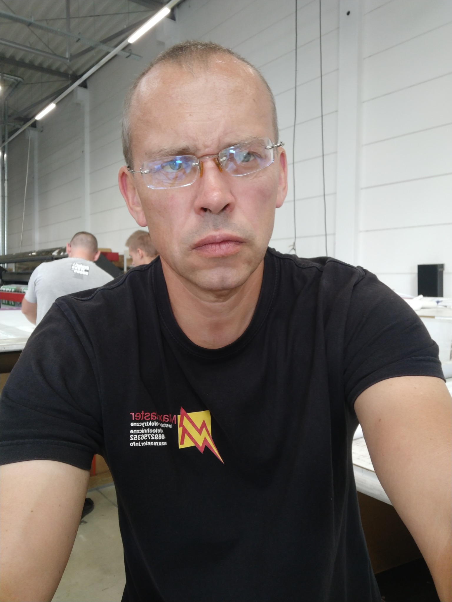 Portret mężczyzny w okularach i czarnym T-shircie z logo firmy, z widocznymi pracownikami w tle w jasnym pomieszczeniu.