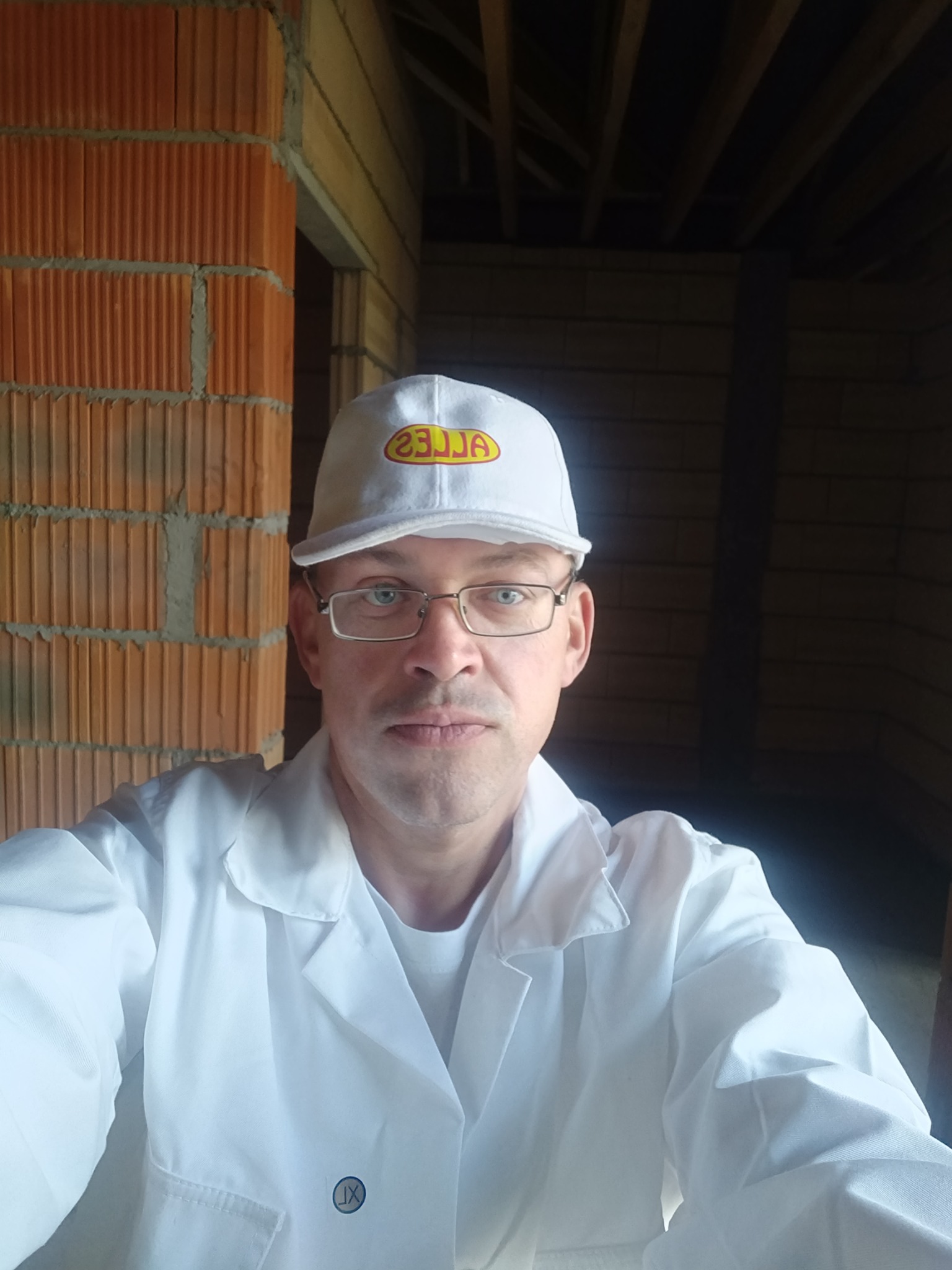 Mężczyzna w okularach i białym uniformie na tle surowych murów z cegły dziurawki i drewnianej konstrukcji stropu w trakcie budowy.