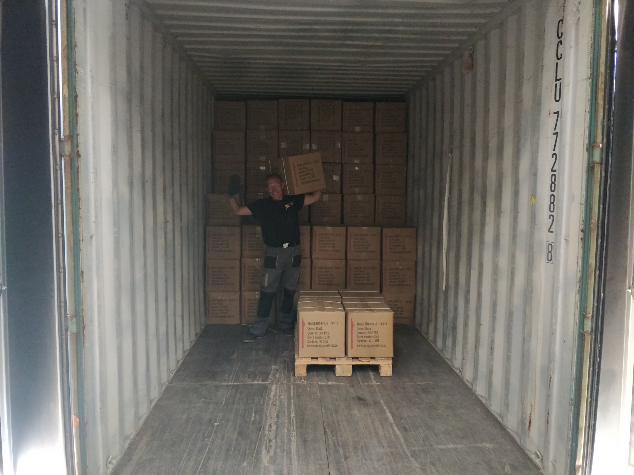 Mężczyzna w roboczym ubraniu i rękawicach unosi karton wewnątrz kontenera transportowego, za nim stosy kartonów ułożonych do sufitu, a na pierwszym planie paleta z dwoma kartonami.