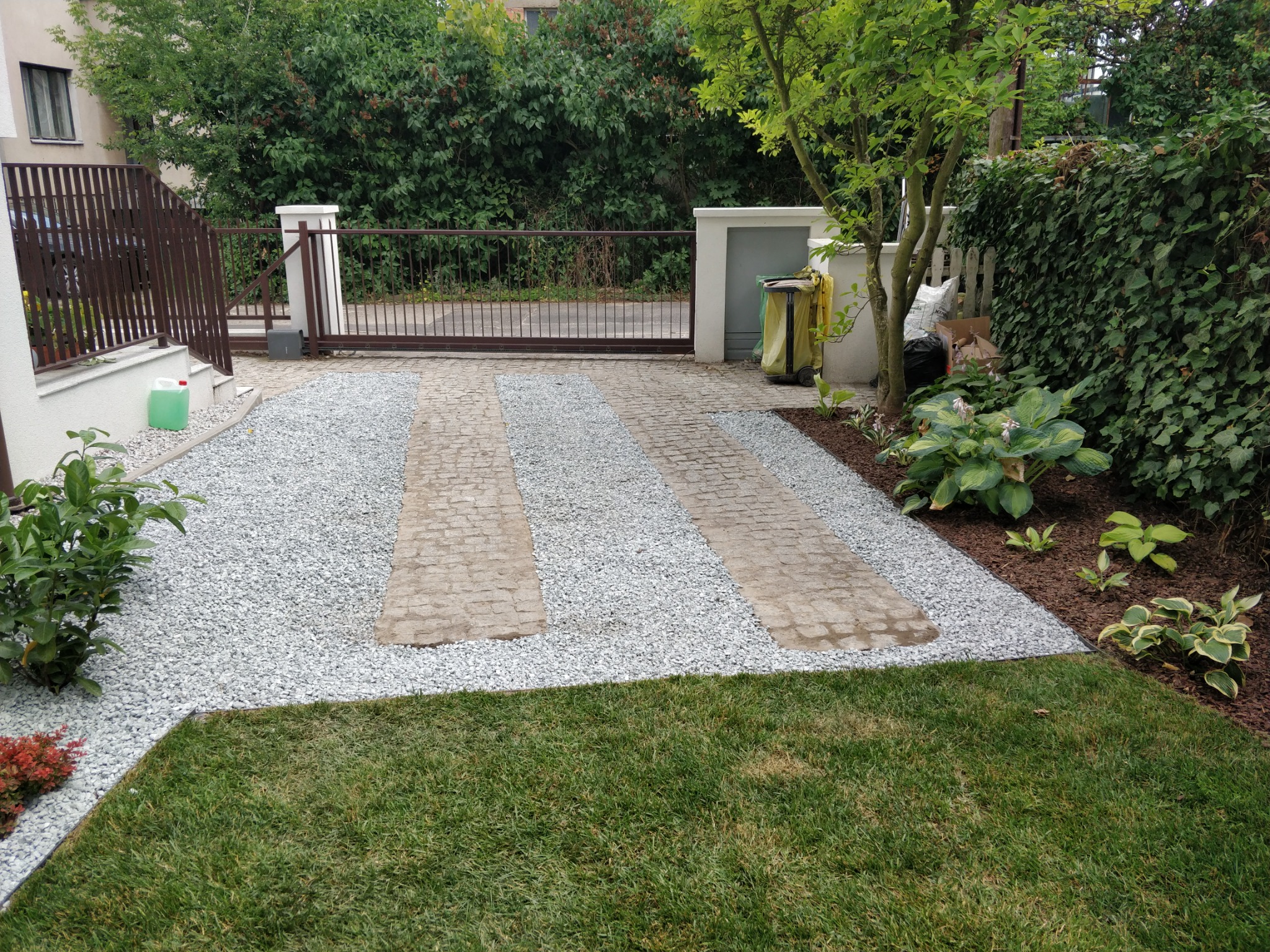 Ułożony podjazd z kostki brukowej i białego grysu, obok trawnik i rabata z roślinami, brama wjazdowa w tle.