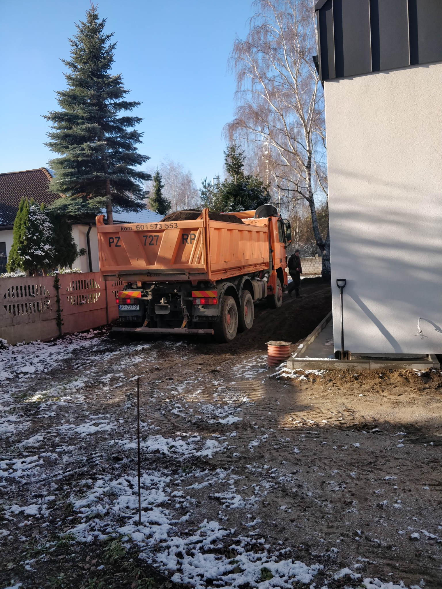 Pomarańczowa wywrotka z ziemią na posesji, częściowo pokrytej śniegiem, w tle dom z zielonym świerkiem, obok nowy budynek z łopatą opartą o ścianę.