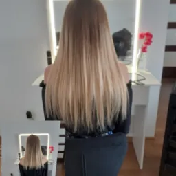 Długie włosy ombre, od brązu u nasady do jasnego blondu na końcach, widok z tyłu, oświetlone lustro w tle, klientka siedzi na czarnym krześle.