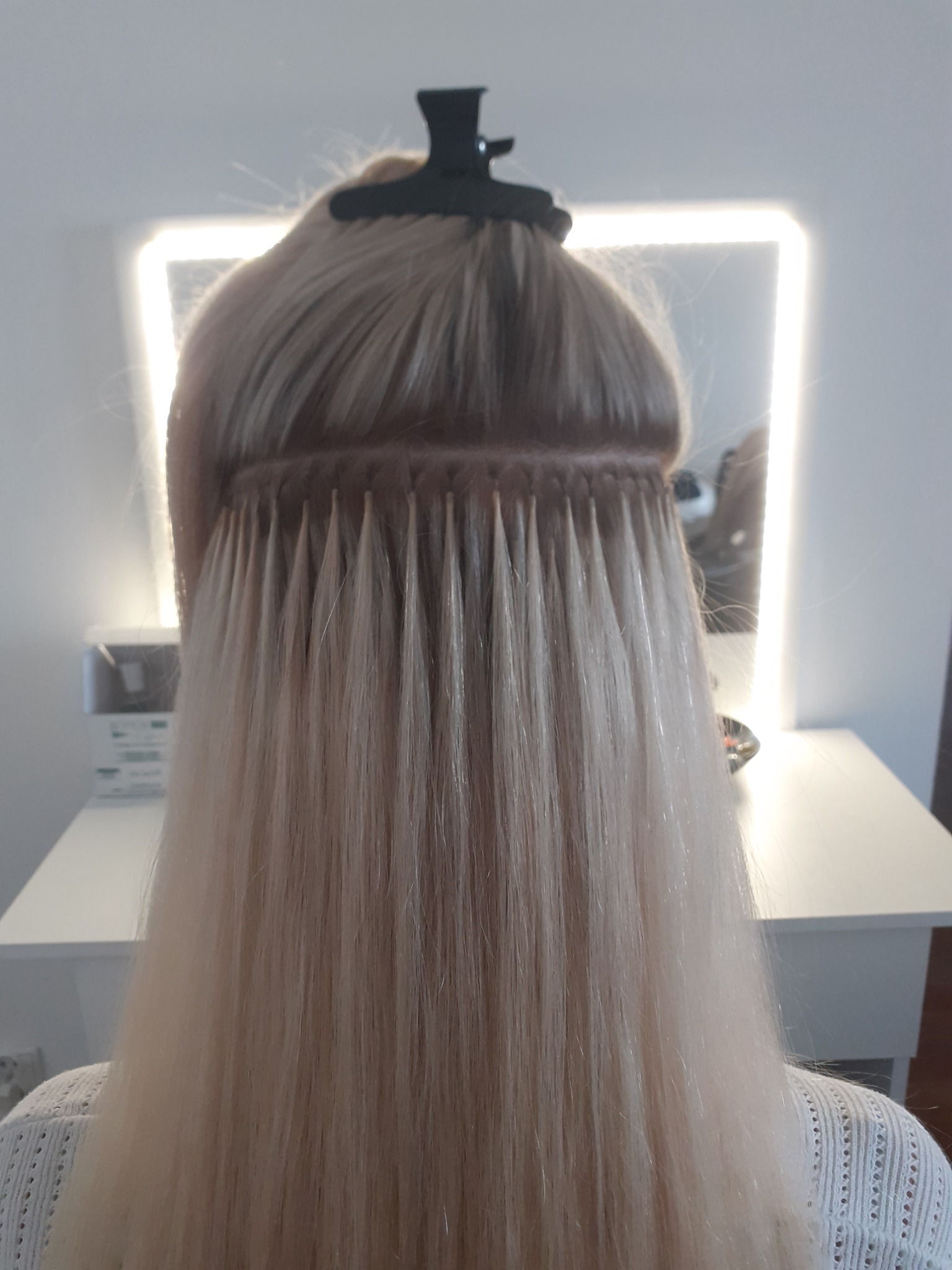 Proces przedłużania włosów metodą keratynową na jasnych włosach, widoczne łączenia, czarny klips przytrzymujący włosy u góry głowy, tło stanowi biała ściana i podświetlane lustro.