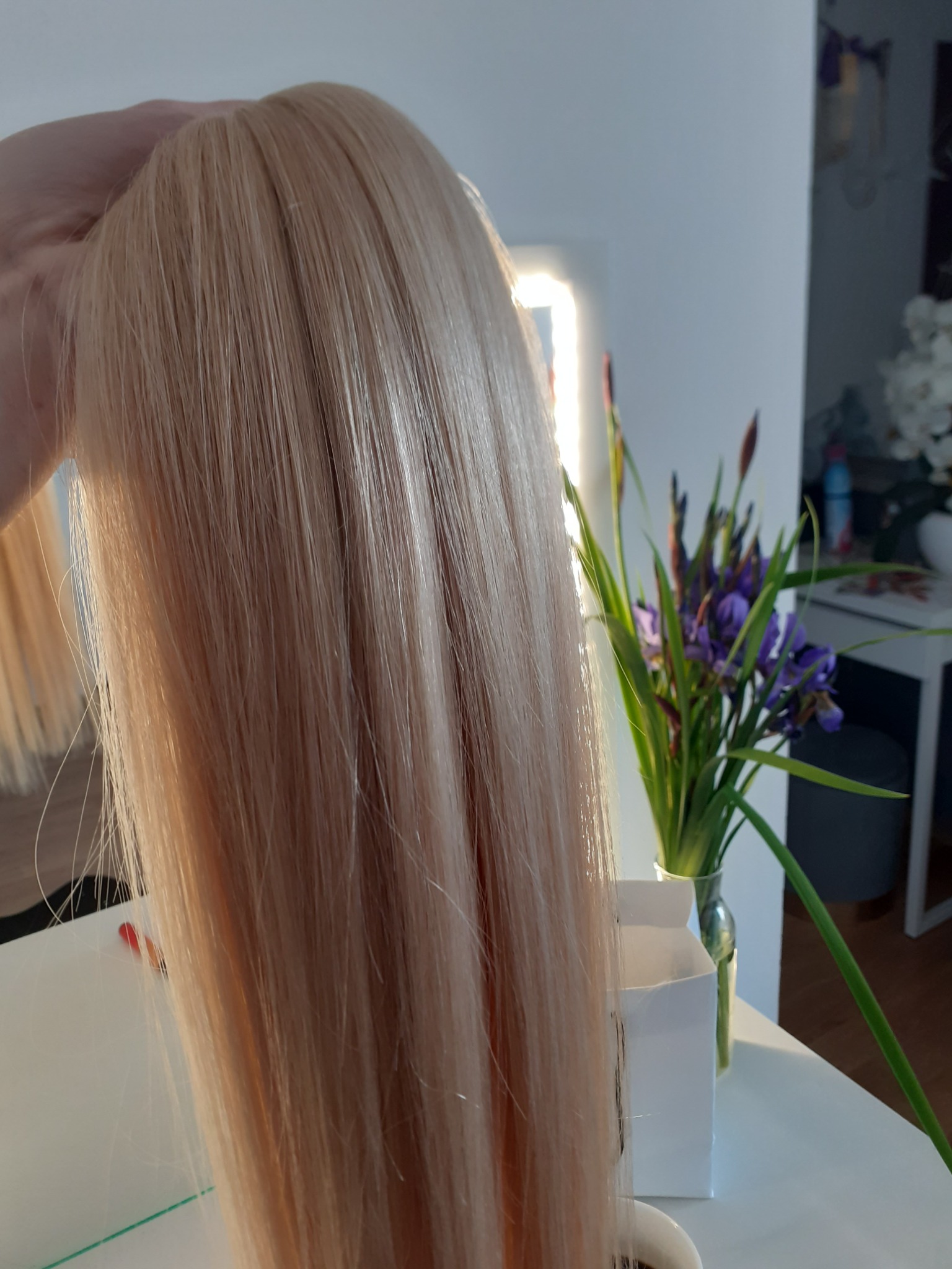 Blond, proste włosy doczepiane trzymane w dłoni, w tle bukiet irysów i fragment białego stolika.