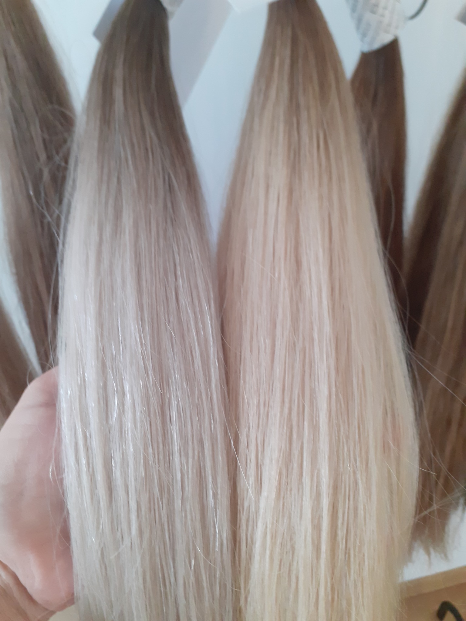 Próbki włosów w różnych odcieniach blondu i brązu, zawieszone na wieszakach, trzymane w dłoni.