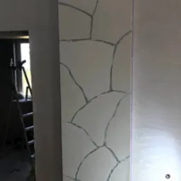 Ściana w trakcie wykańczania z efektem kamiennej mozaiki; szpachlowana powierzchnia z widocznymi liniami imitującymi kamienne płyty, drabina w tle, pomieszczenie w trakcie prac wykończeniowych.