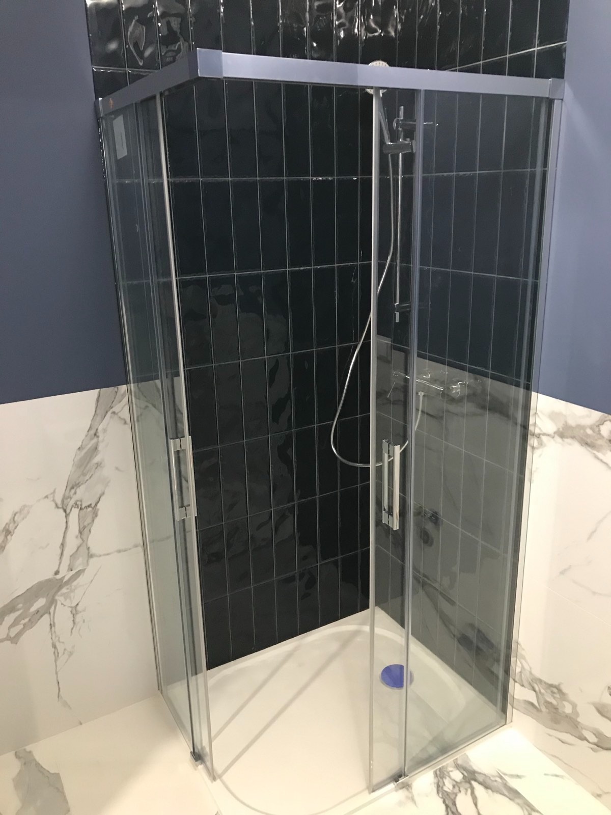 Narożna kabina prysznicowa z przezroczystego szkła z chromowanymi profilami, na białym brodziku z niebieskim elementem odpływowym, na tle ściany z czarnych, błyszczących płytek ułożonych pionowo...