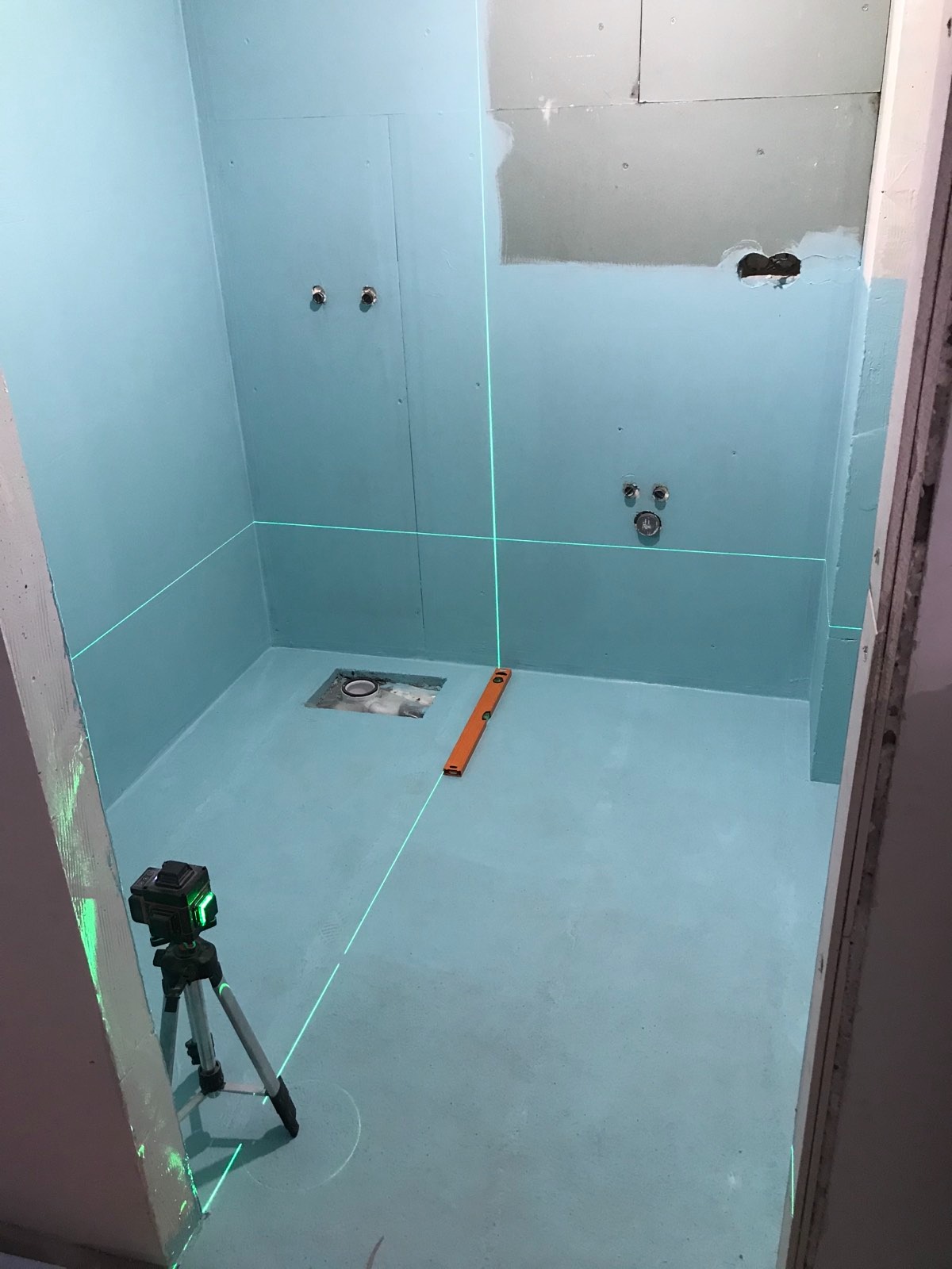 Przygotowanie łazienki do dalszych prac wykończeniowych: ściany i podłoga pokryte hydroizolacją w odcieniach błękitu, poziomica laserowa wyznacza pion i poziom, widoczne przyłącza...