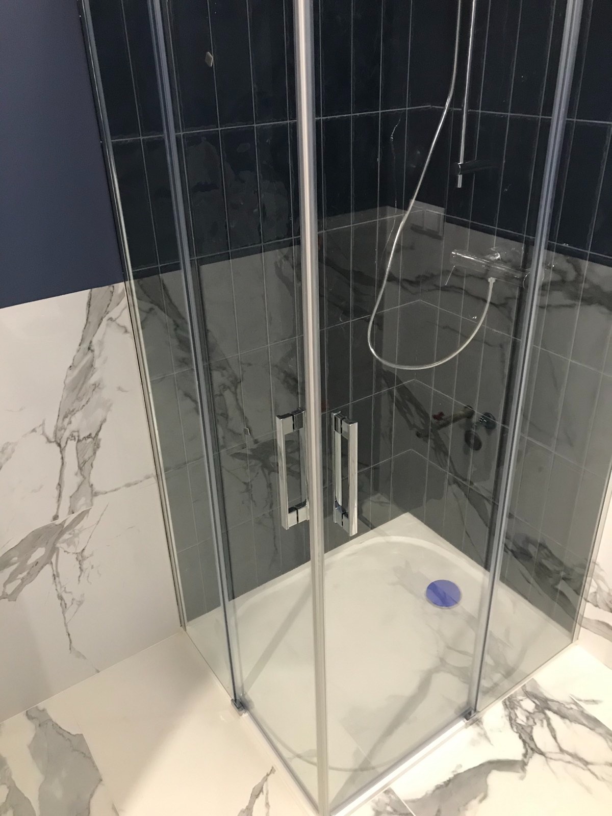 Nowoczesna kabina prysznicowa z przezroczystego szkła, chromowanymi uchwytami i brodzikiem z niebieskim odpływem, otoczona płytkami imitującymi marmur i ciemnymi, pionowymi płytkami.