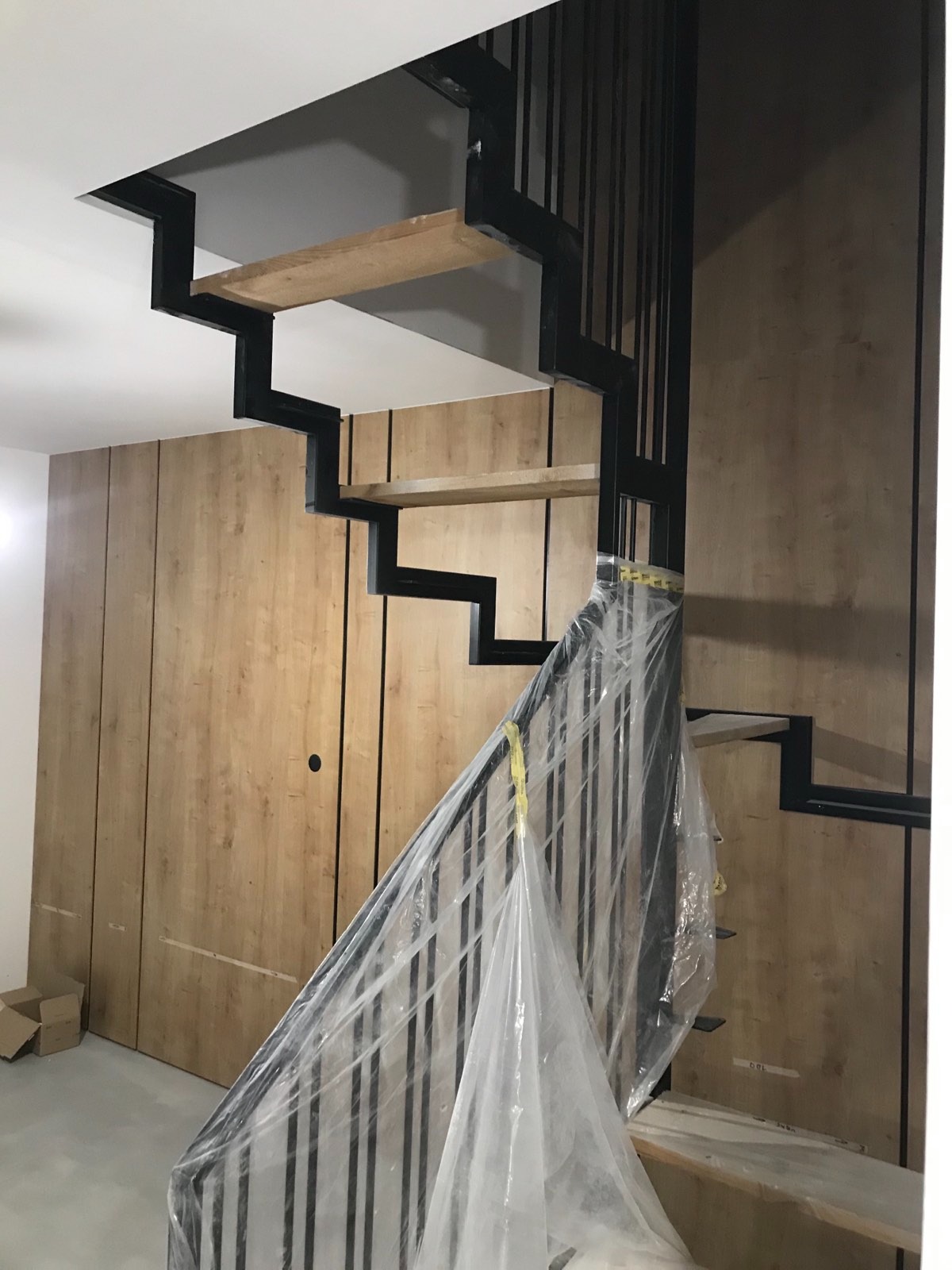 Nowoczesne schody wiszące z czarną metalową konstrukcją i drewnianymi stopniami, częściowo zasłonięte folią ochronną, na tle ściany wykończonej drewnopodobnymi panelami.