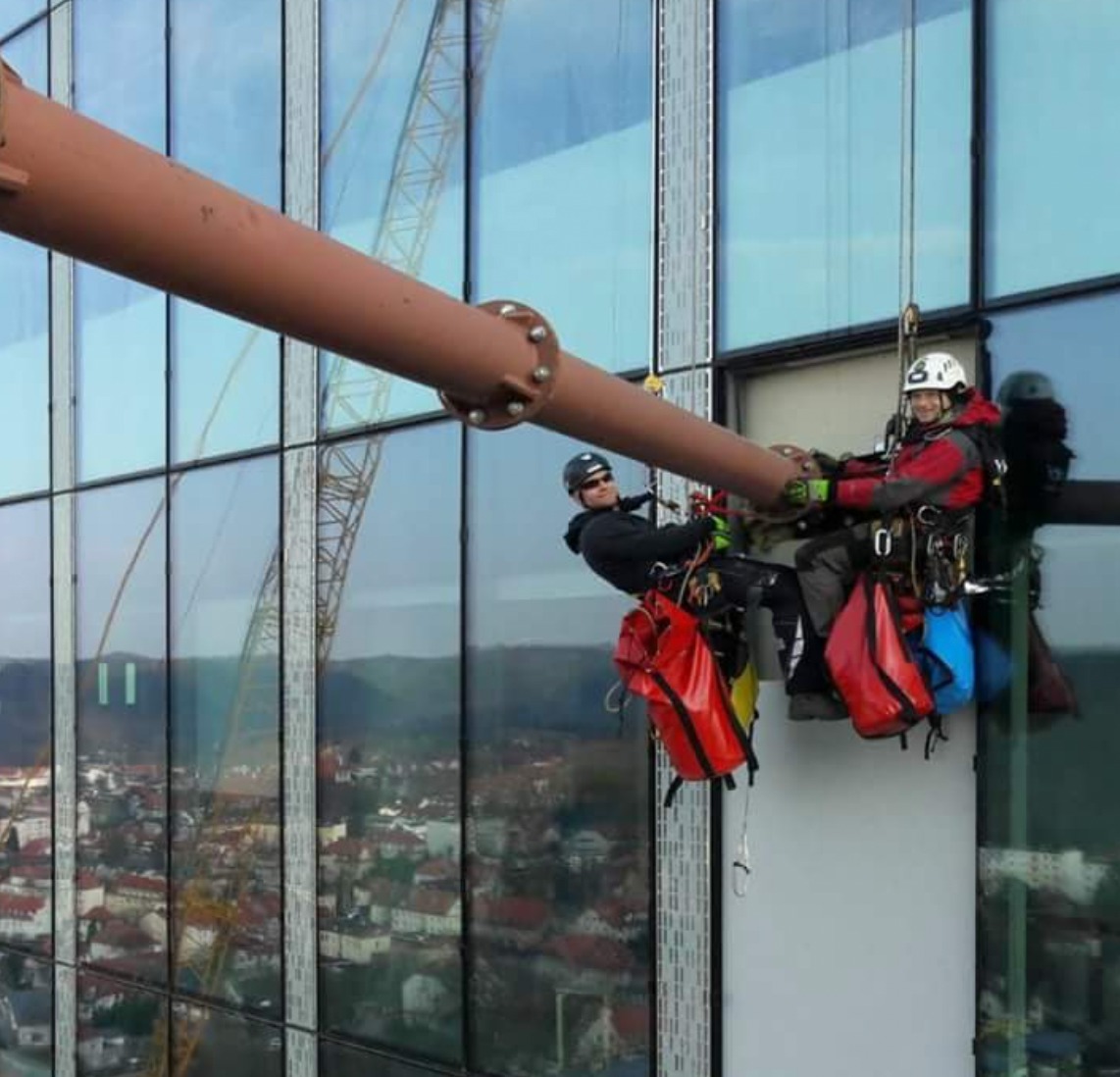 Dwóch alpinistów przemysłowych w uprzężach, pracujących na zewnątrz szklanego budynku, wspierających się na dużej metalowej rurze, widok na miasto w tle.