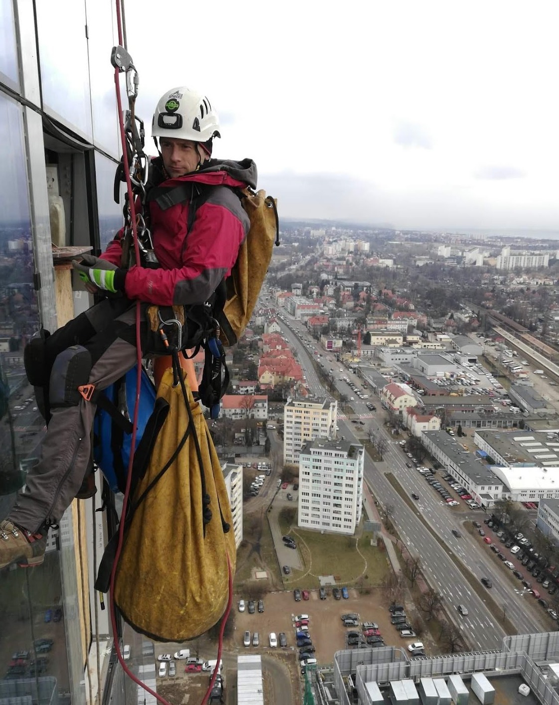 Pracownik w kasku, zabezpieczony linami, wykonuje prace na wysokości na szklanej fasadzie budynku, widok na miasto w tle.