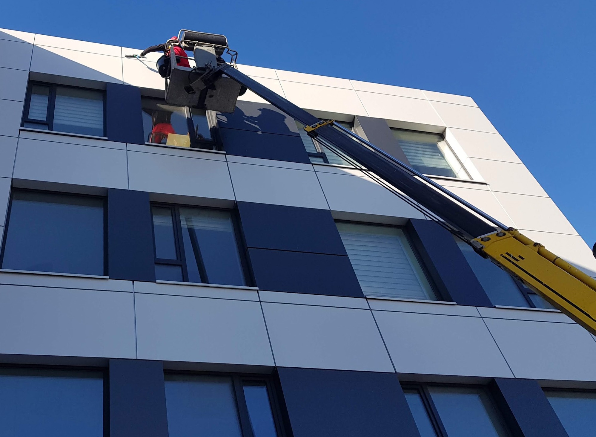 Pracownik w czerwonym kombinezonie, znajdujący się w podnośniku koszowym, czyści okna na elewacji nowoczesnego biurowca o geometrycznym wzorze z paneli w kolorze białym i grafitowym.