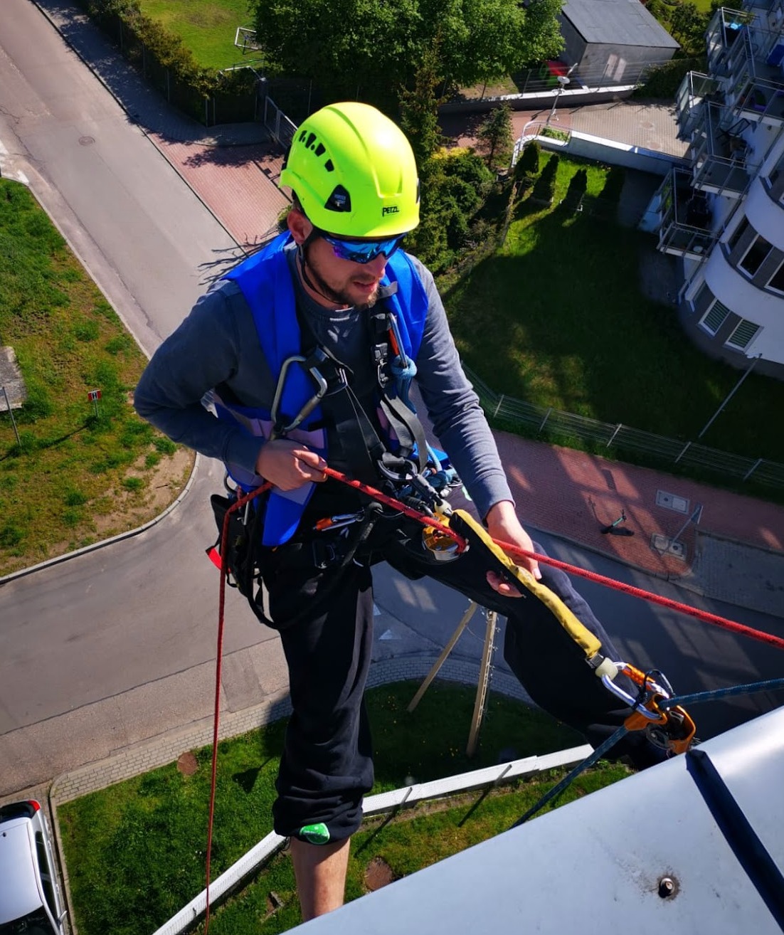 Pracownik w kasku i uprzęży, z perspektywy dachu, przygotowuje się do pracy na wysokości, widoczne liny, karabinki i elementy zabezpieczające, w tle budynek mieszkalny i zieleń.