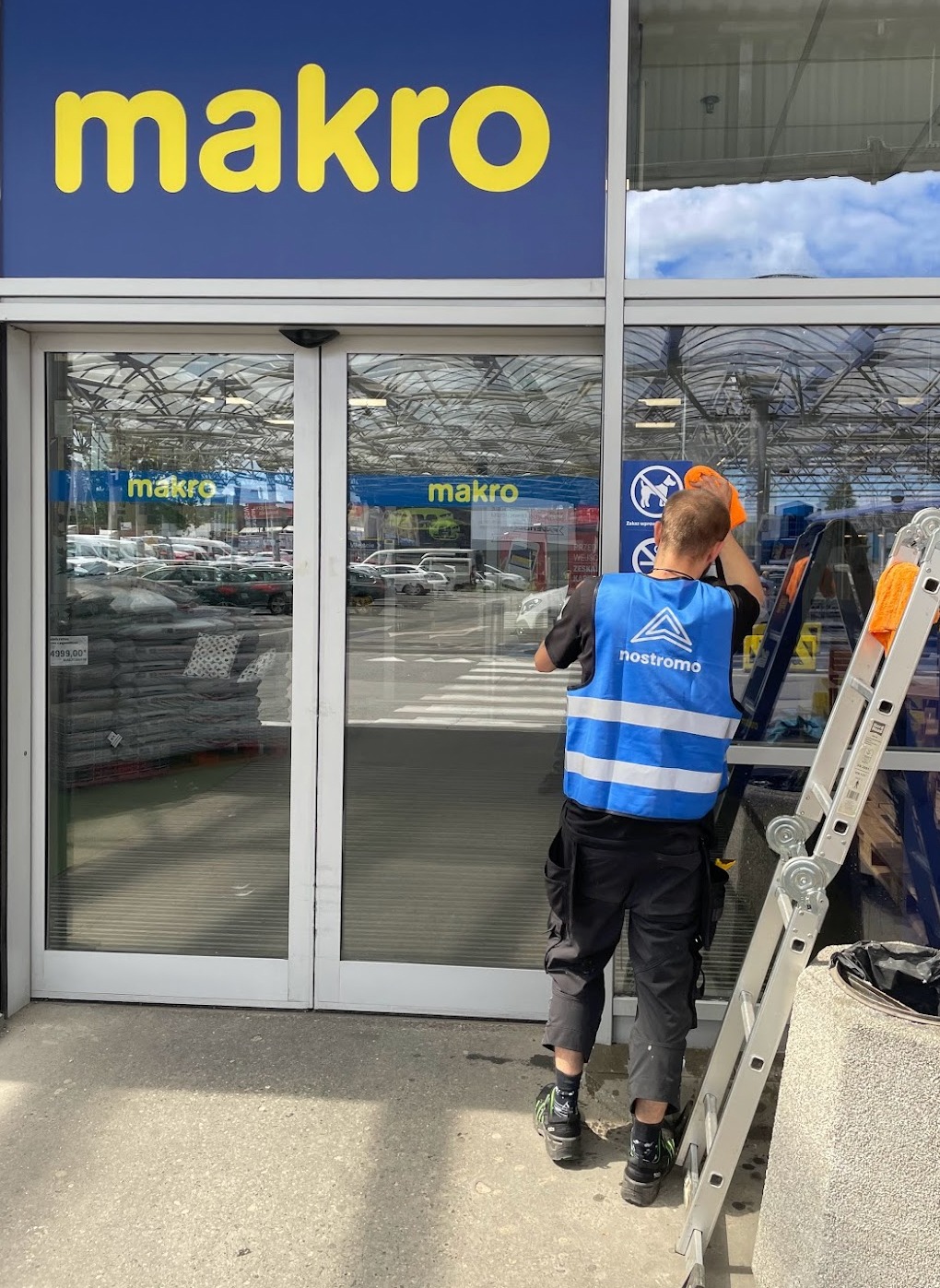 Pracownik w kamizelce odblaskowej z logo firmy czyści szklane drzwi wejściowe do sklepu Makro, używając pomarańczowej ściereczki i drabiny.