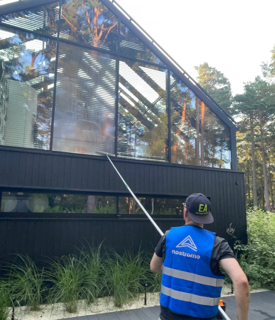 Mężczyzna w kamizelce odblaskowej myje okna nowoczesnego domu z dużymi przeszkleniami przy użyciu długiego kija z myjką. Widoczne odbicie drzew w oknach.