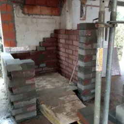 Wnętrze w trakcie budowy w Kałuszynie, widoczne fragmenty ścian z czerwonej cegły i szarych bloczków, drewniane deski podłogowe, metalowe podpory stropowe, surowy stan wykończenia.