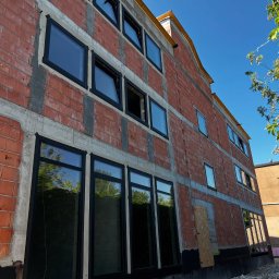 Aluzone - Budynek w trakcie budowy z zamontowanymi czarnymi oknami. Widoczne cegły, betonowe wzmocnienia i fragment nieba. Ujęcie z dołu, podkreślające wysokość budynku.