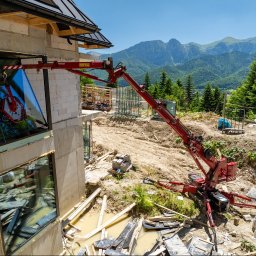 Aluzone - Montaż okna na budowie apartamentów w Zakopanem. Czerwony dźwig podnosi szybę, w tle góry i las. Widoczne materiały budowlane i elementy konstrukcyjne.