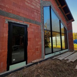 Aluzone - Surowy stan domu w stylu stodoły z zamontowanymi oknami o nietypowym, trójkątnym kształcie i czarnych ramach. Widoczne detale konstrukcyjne i materiały budowlane.