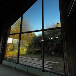 Aluzone - Wysokie okno z czarnymi szprosami w surowym wnętrzu domu w stylu stodoły. Widok na zieleń i błękitne niebo. Nietypowy kształt okna.