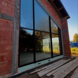 Aluzone - Duże, trójkątne okno z czarną ramą w surowej ścianie z cegły dziurawki, widoczne deski i pianka montażowa. Dom w stylu stodoły w trakcie budowy.