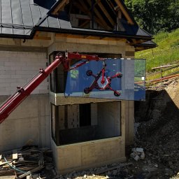 Aluzone - Montaż okna w surowym budynku z użyciem dźwigu ssącego. Widoczni pracownicy w odbiciu szyby. Budowa apartamentów w Zakopanem w tle.