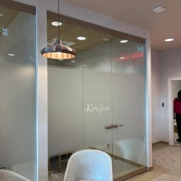 Aluzone - Szklana ścianka działowa z napisem 'Krajny' w biurze PEKOSA, oświetlona wiszącą lampą. Widoczne krzesła i fragment korytarza.