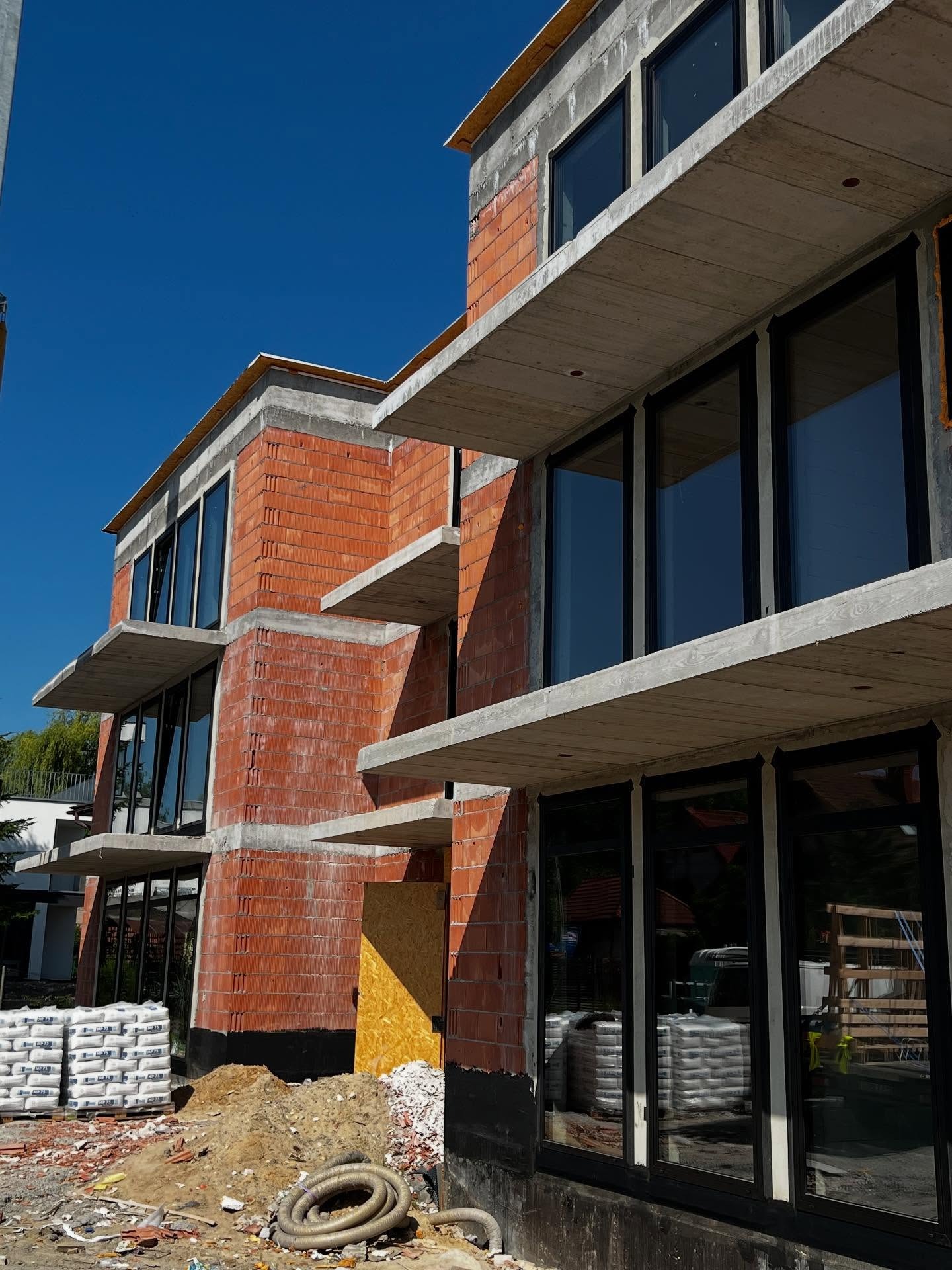 Budynek usługowy w trakcie budowy z widocznymi oknami z czarnymi ramami, ceglaną elewacją i betonowymi elementami konstrukcyjnymi. Na dole worki z zaprawą.