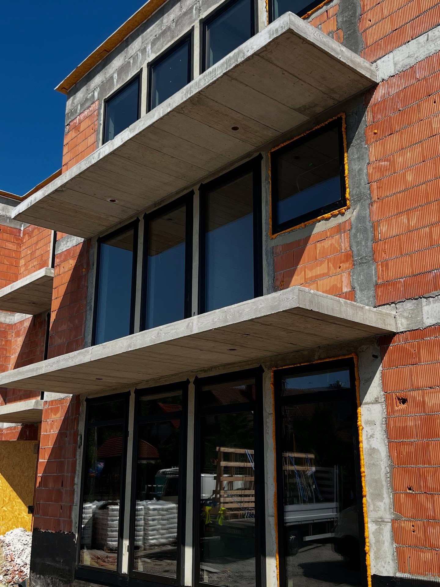 Budynek w trakcie budowy z zamontowanymi oknami z czarnymi ramami i betonowymi balkonami. Widoczne cegły i pianka montażowa wokół okien. Słoneczny dzień.