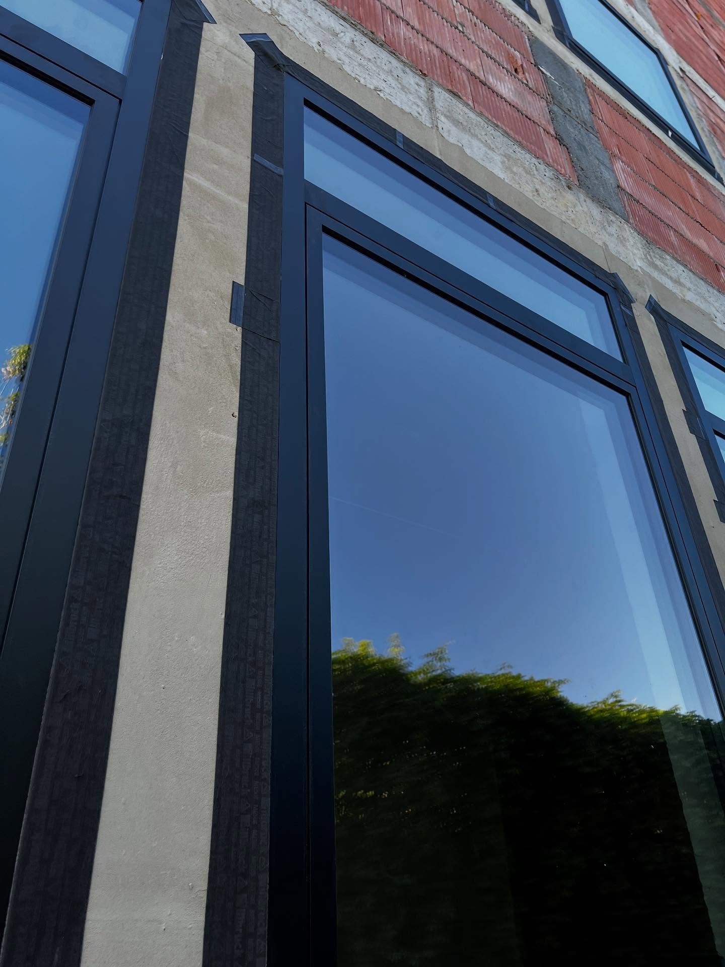 Montaż okien w budynku usługowym: widok z dołu na czarne ramy okienne, surowy mur z cegły i beton, zabezpieczenia montażowe. Odbicie zieleni w szybach.