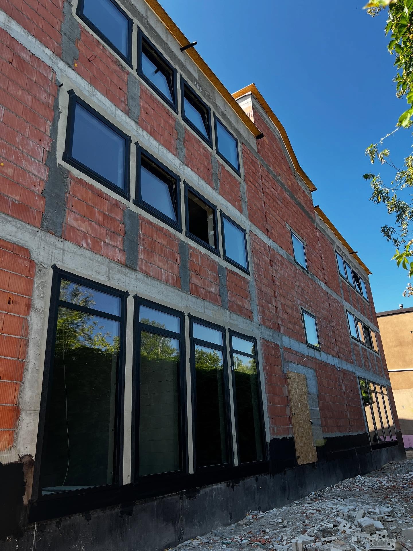 Budynek w trakcie budowy z zamontowanymi czarnymi oknami. Widoczne cegły, betonowe wzmocnienia i fragment nieba. Ujęcie z dołu, podkreślające wysokość budynku.