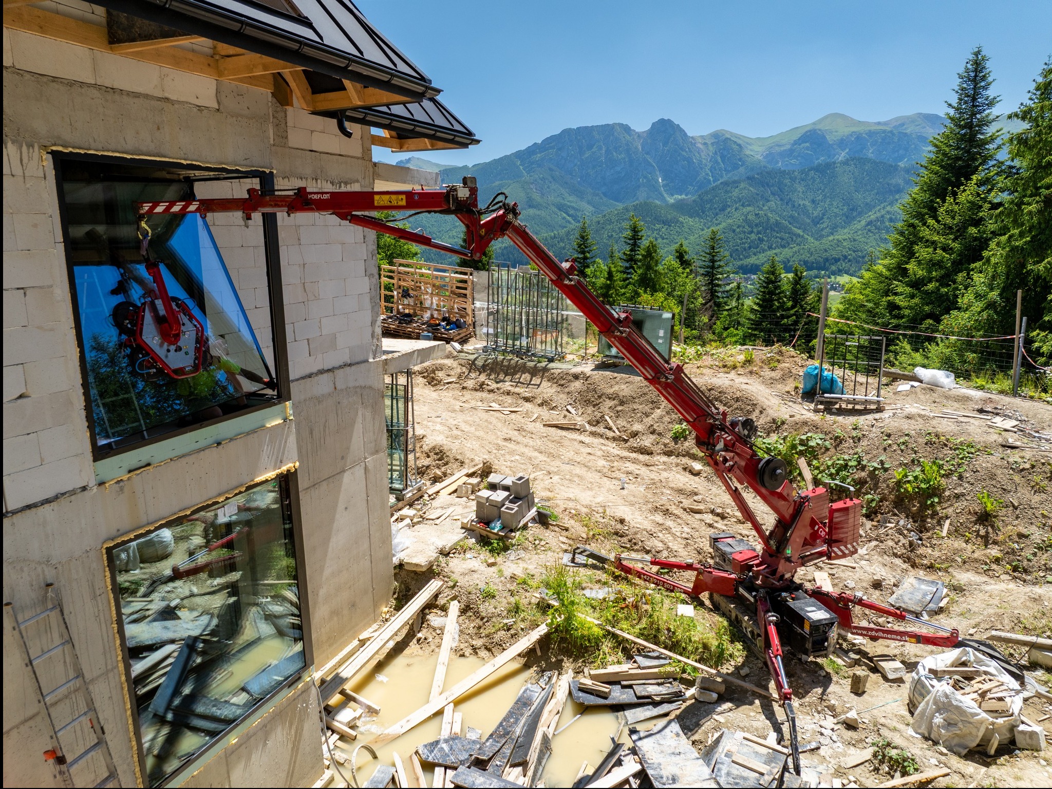 Montaż okna na budowie apartamentów w Zakopanem. Czerwony dźwig podnosi szybę, w tle góry i las. Widoczne materiały budowlane i elementy konstrukcyjne.
