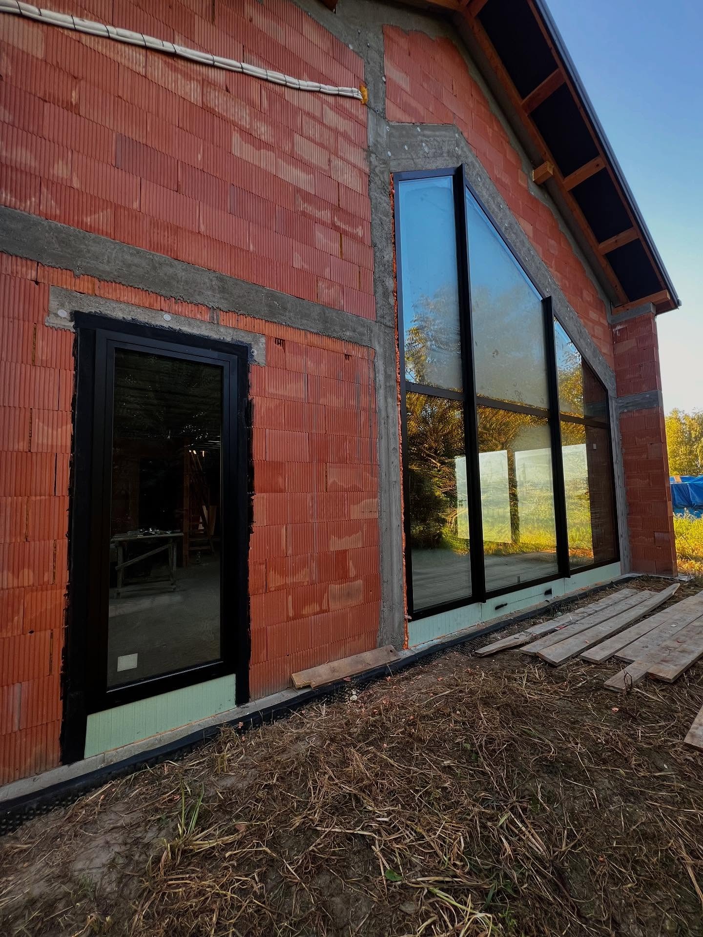 Surowy stan domu w stylu stodoły z zamontowanymi oknami o nietypowym, trójkątnym kształcie i czarnych ramach. Widoczne detale konstrukcyjne i materiały budowlane.