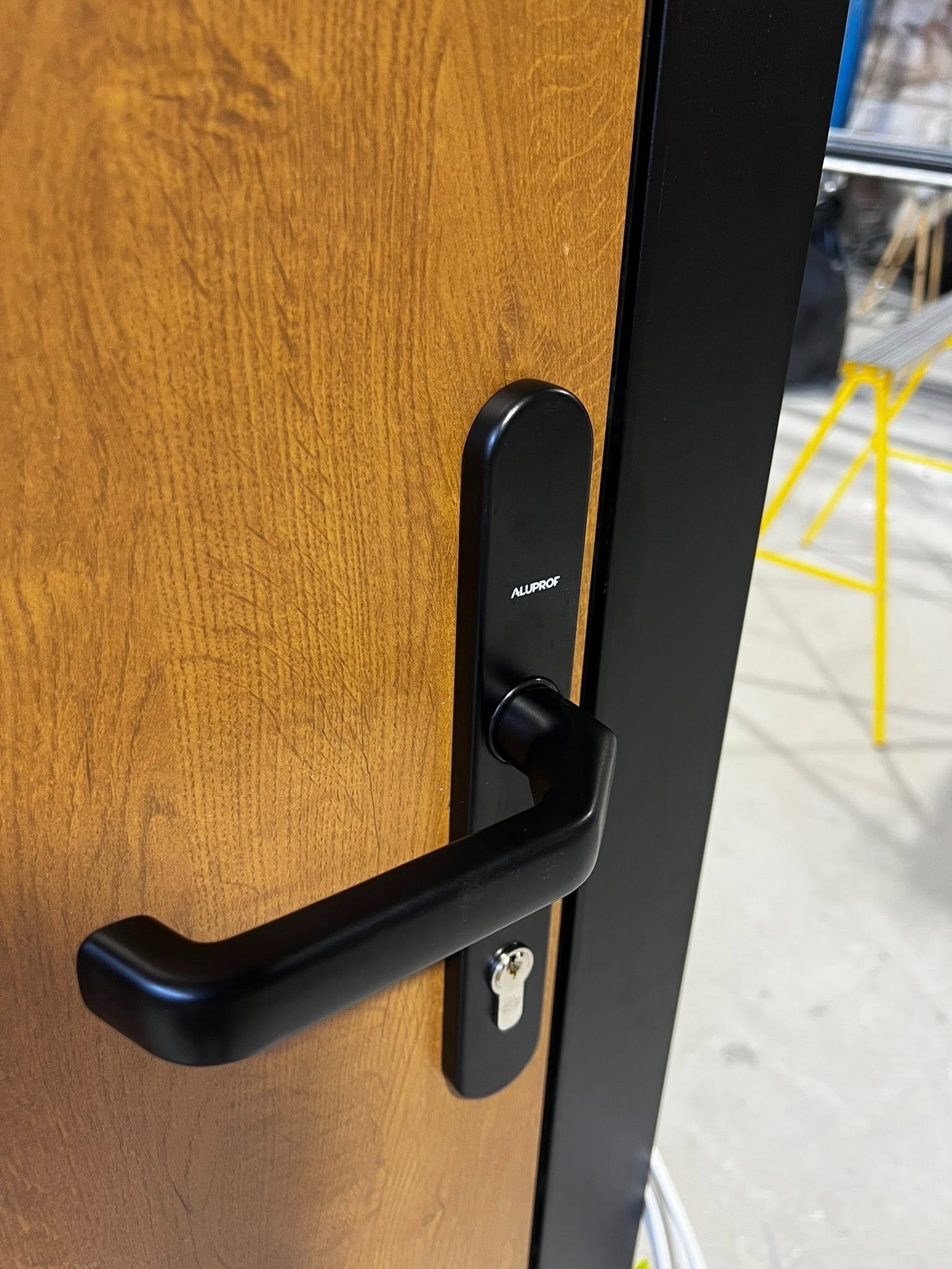 Częściowy widok drzwi z okleiną drewnopodobną, czarną ramą i klamką z szyldem marki Aluprof. W tle widoczne krzesło i elementy wyposażenia.
