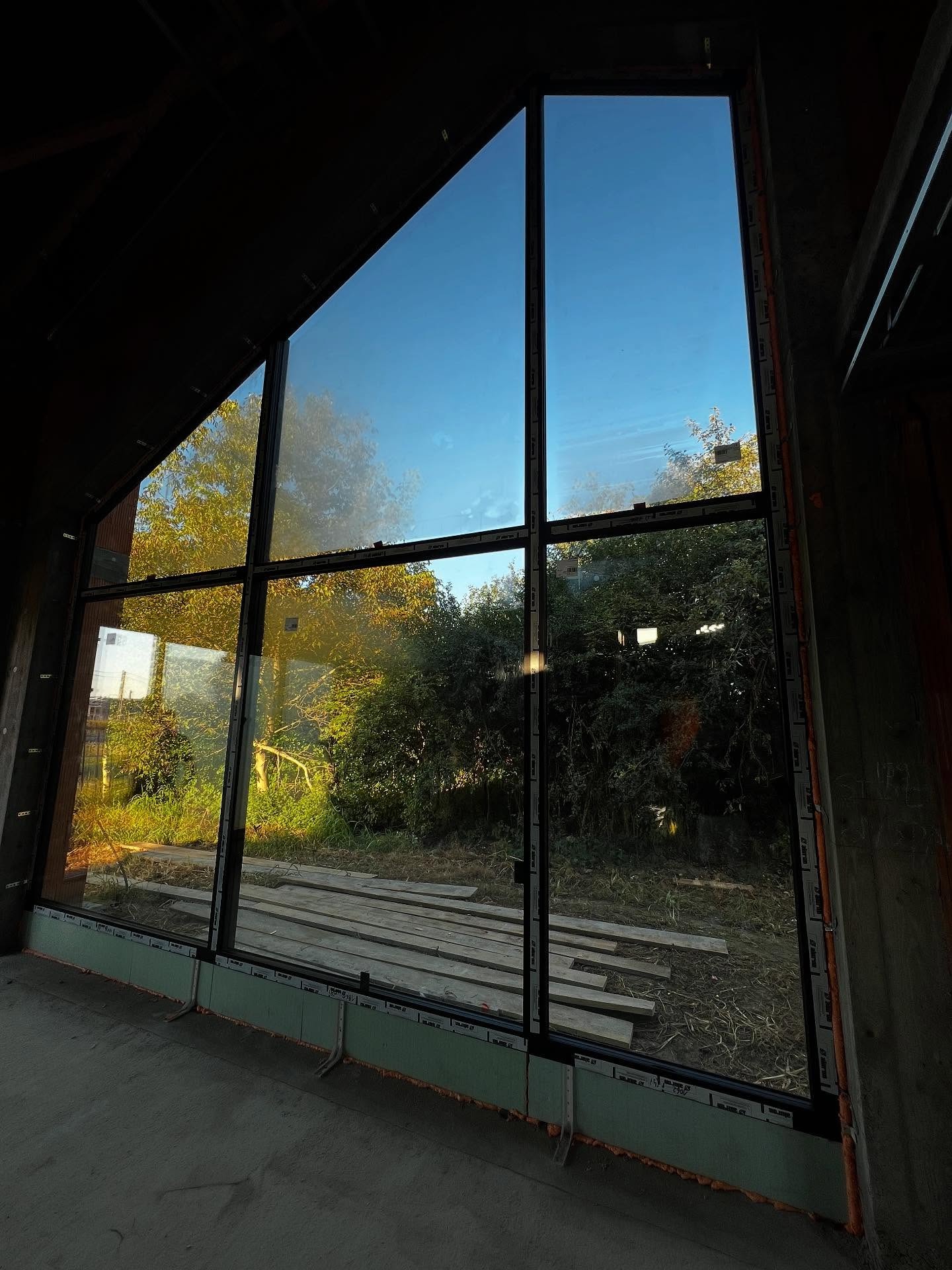 Wysokie okno z czarnymi szprosami w surowym wnętrzu domu w stylu stodoły. Widok na zieleń i błękitne niebo. Nietypowy kształt okna.