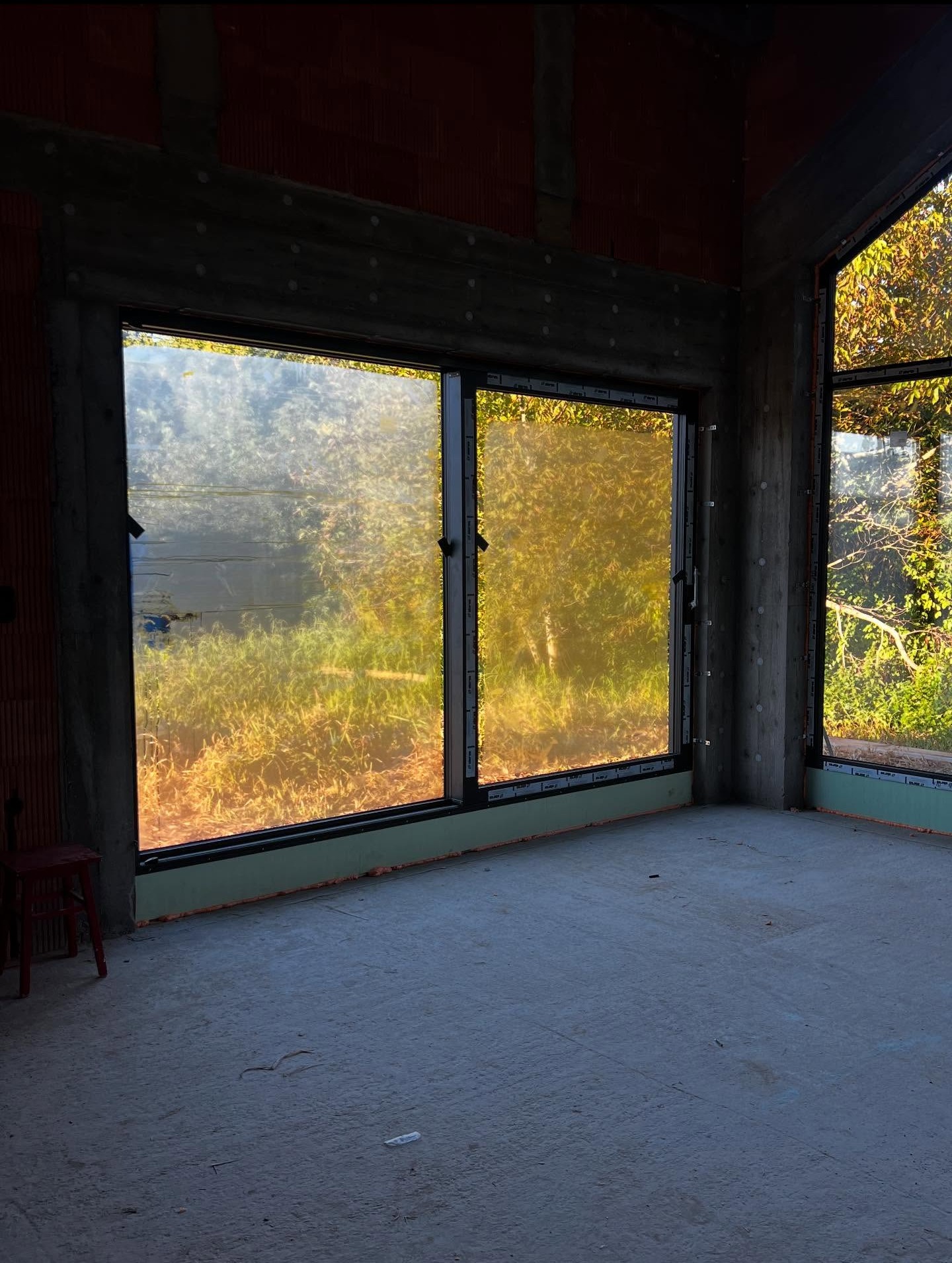 Wnętrze surowego domu w stylu stodoły z dużymi oknami z widokiem na zieleń. Widoczne surowe ściany i betonowa podłoga. W tle zieleń i drzewa.