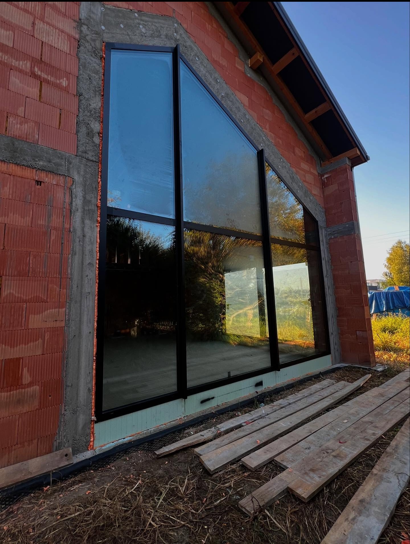 Duże, trójkątne okno z czarną ramą w surowej ścianie z cegły dziurawki, widoczne deski i pianka montażowa. Dom w stylu stodoły w trakcie budowy.