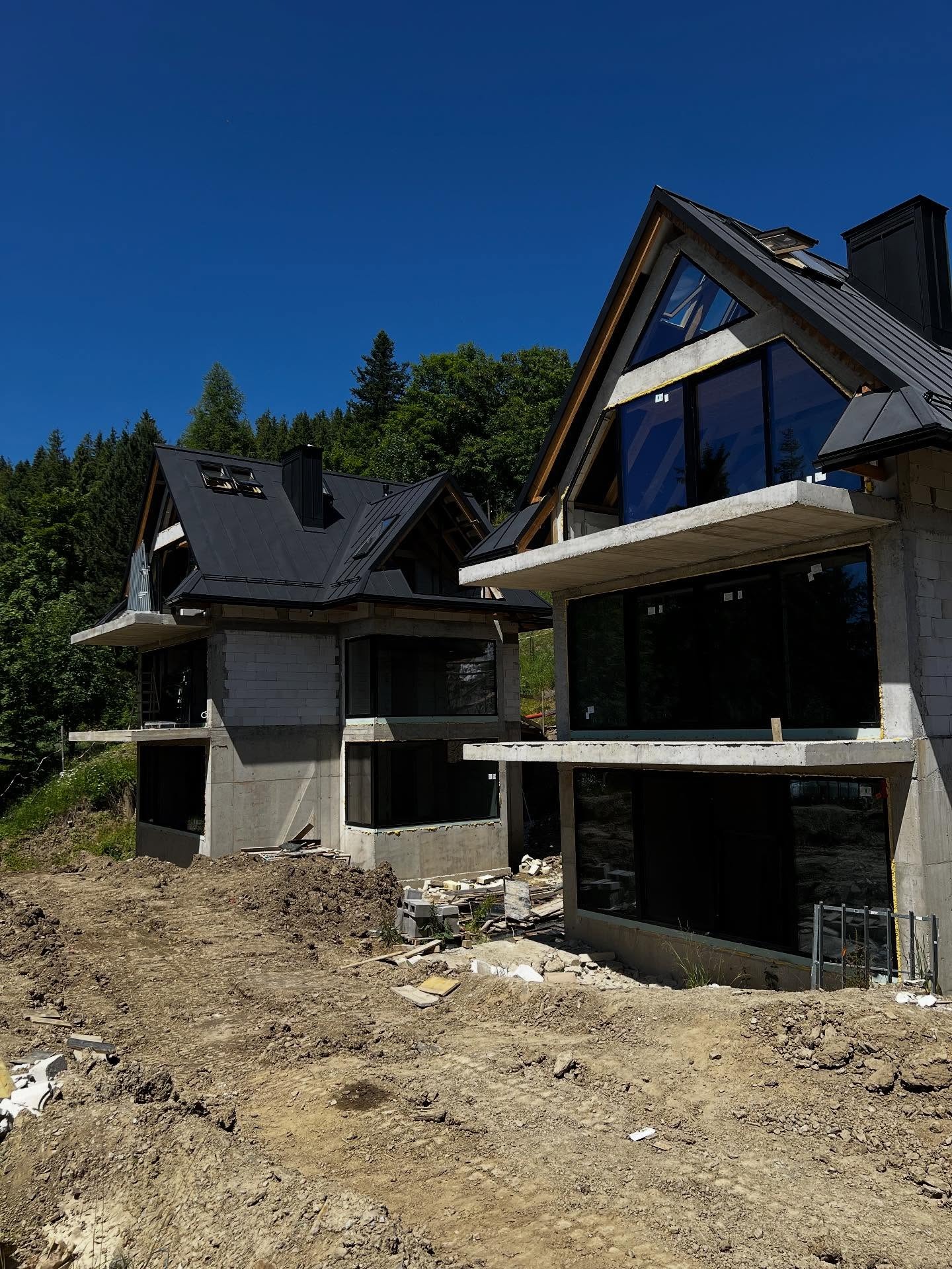 Dwa nowoczesne, surowe apartamenty w stylu górskim w Zakopanem, z dużymi oknami i betonowymi elementami, w trakcie budowy, na tle lasu i błękitnego nieba.