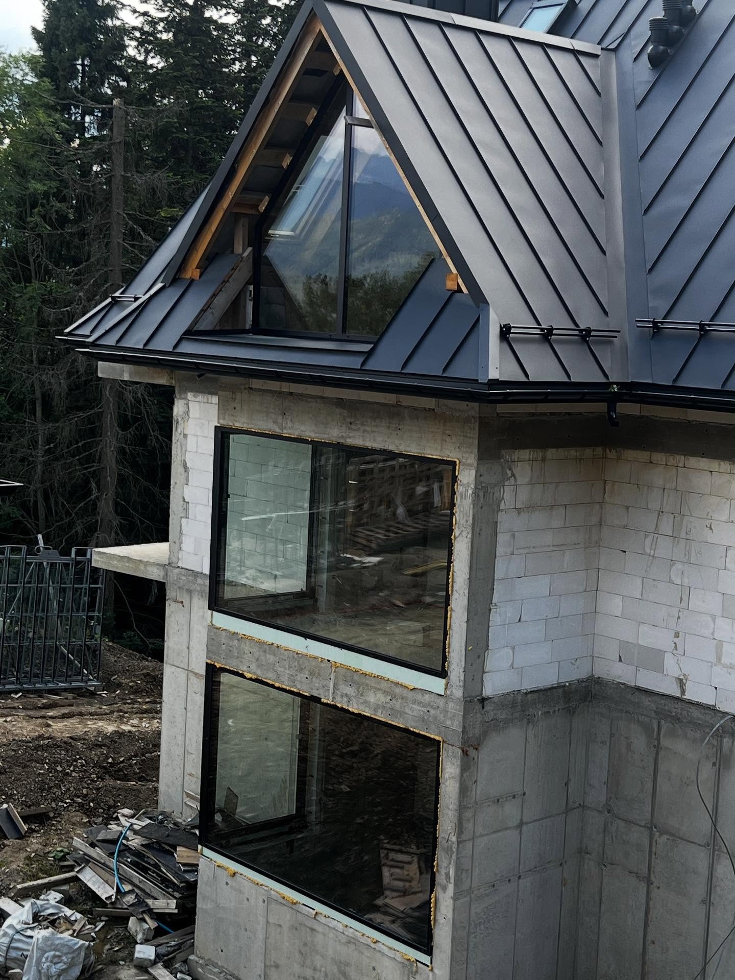 Surowy stan apartamentu w Zakopanem: czarne ramy okien w betonie, skośny dach z blachy na tle lasu i gór. Widoczne materiały budowlane.