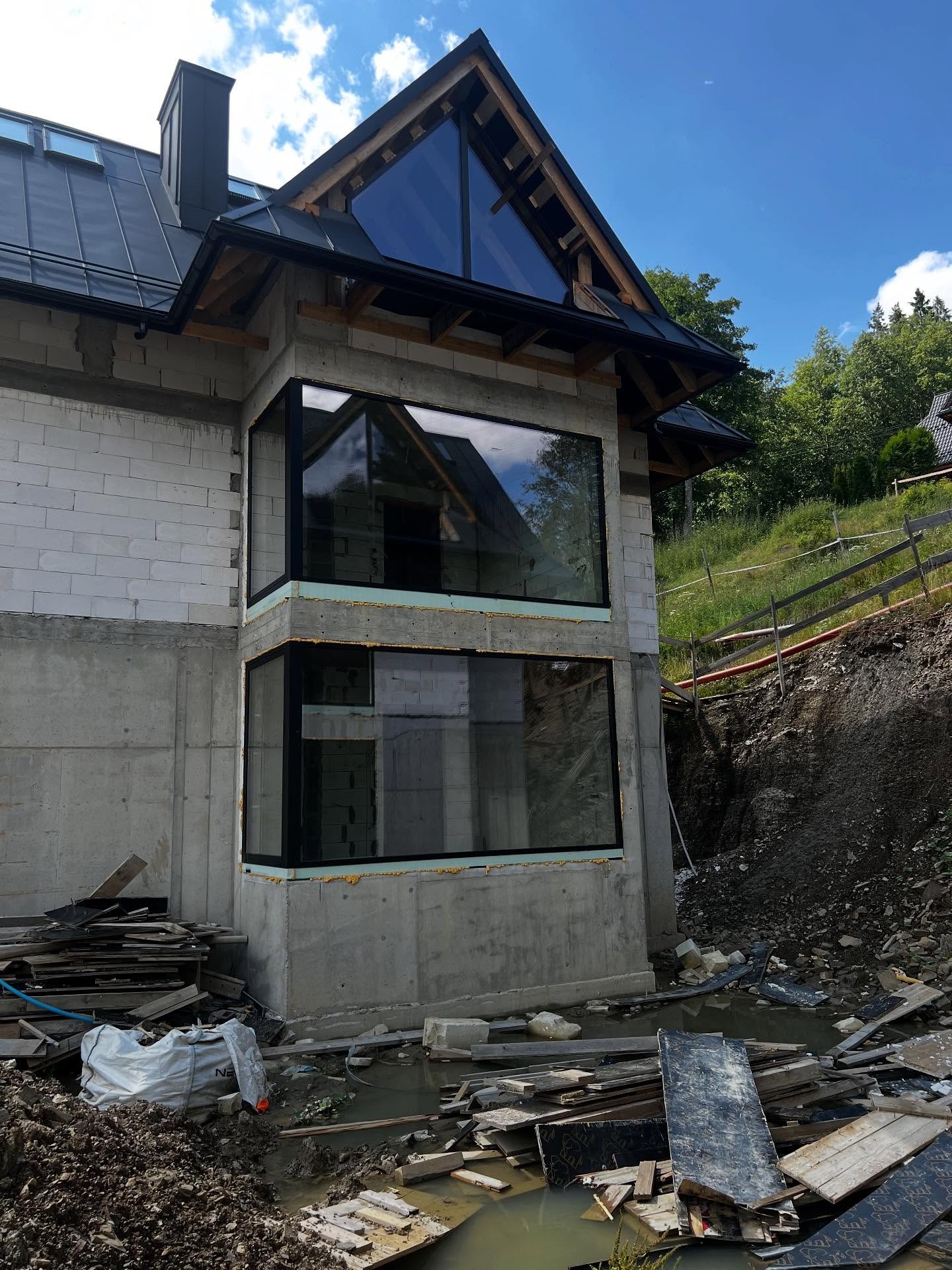 Surowy stan budynku z zamontowanymi oknami z czarnymi ramami. Widoczne materiały budowlane i teren w trakcie prac ziemnych. Okna odbijają krajobraz.