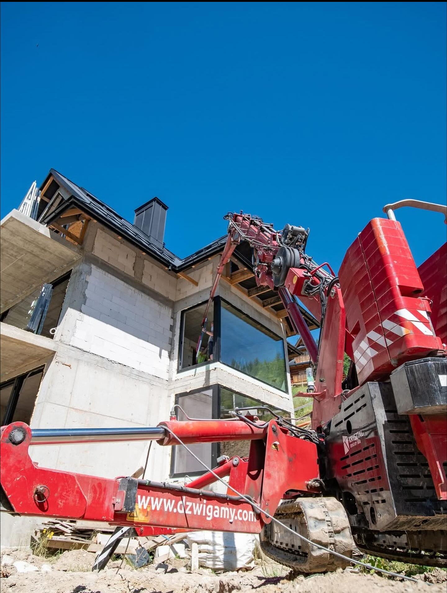 Montaż okien w apartamentach Zakopane z użyciem czerwonego dźwigu. Widok na budynek w stanie surowym otwartym i okna z czarnymi ramami.