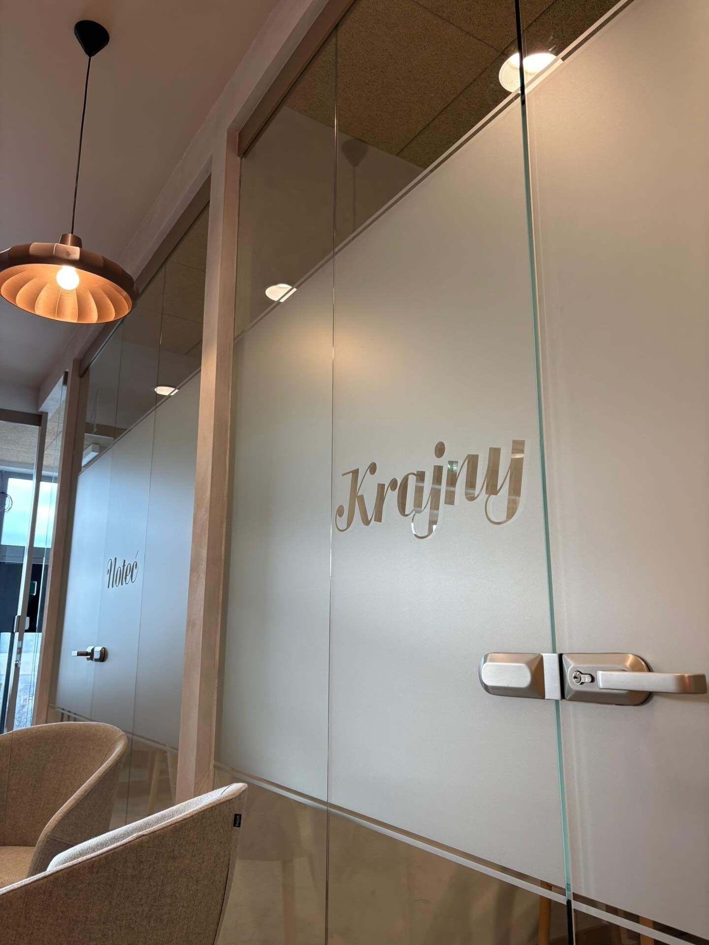 Szklana ścianka działowa z matową folią i napisem 'Krajny' w biurze, widoczny fragment fotela i lampa sufitowa. Nowoczesny design.