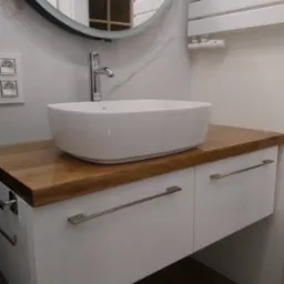 Nowoczesna łazienka z białą umywalką nablatową, drewnianym blatem i szafką z podłużnymi uchwytami, okrągłe lustro z czarną ramą i grzejnik drabinkowy na ścianie z marmurowym wzorem.